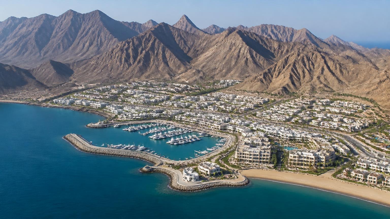 Luchtfoto van een kustgemeenschap in Oman met witte woningen rond een marina en bergen op de achtergrond
