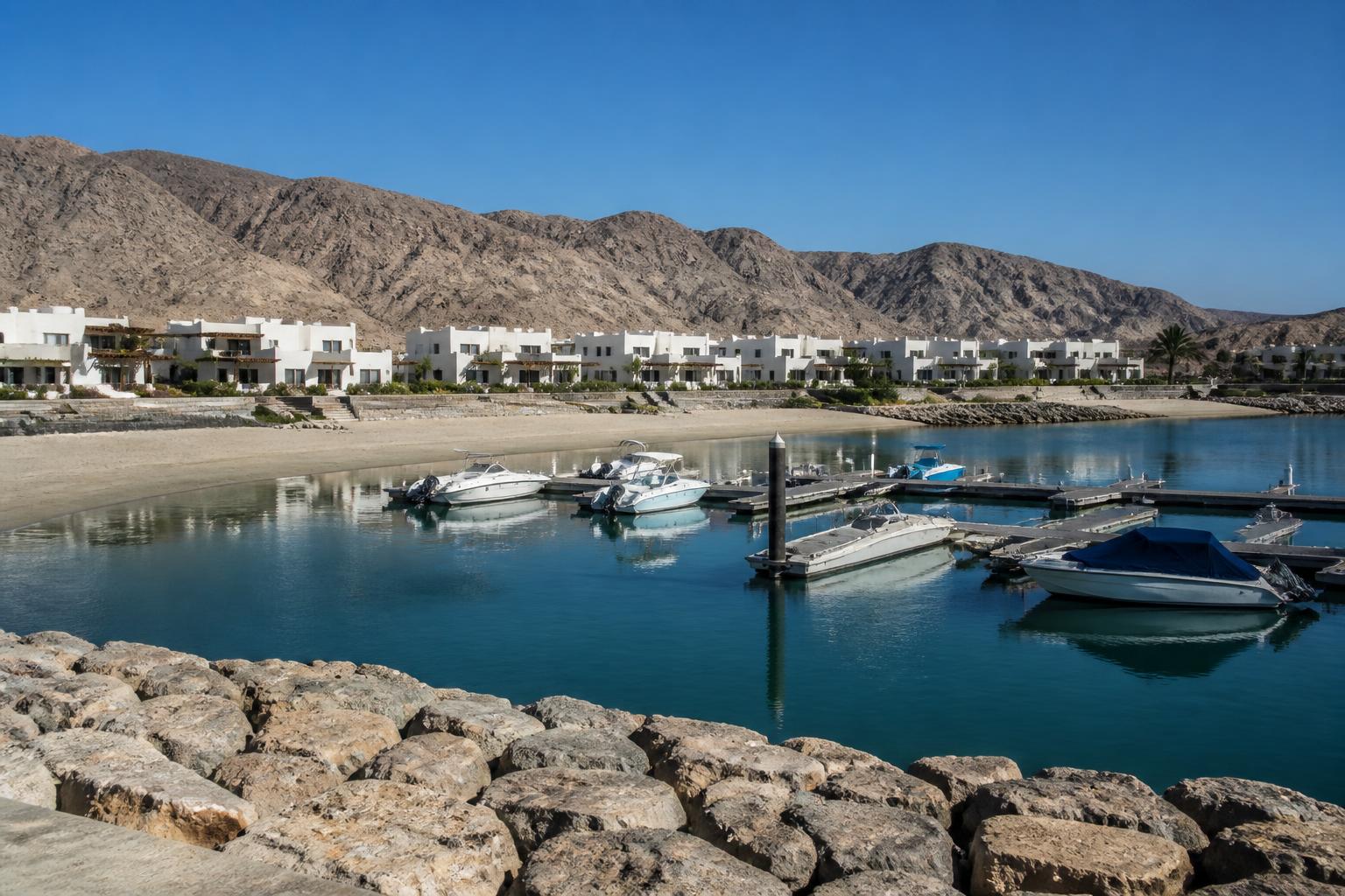 Witte lage woningen aan een rustige marina in Jebel Sifah met boten en kale bergen op de achtergrond