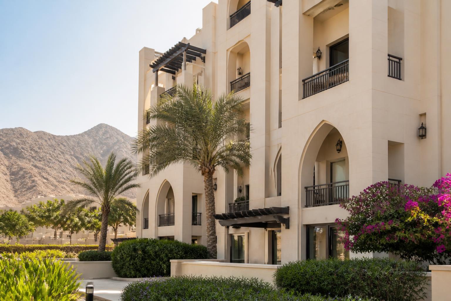 Appartementengebouw in Muscat met crèmekleurige gevel, boogvormige balkons en palmen tegen droge bergen